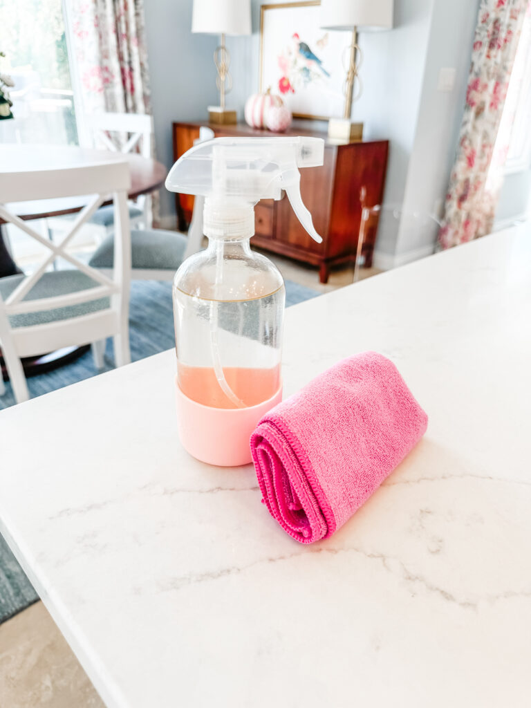 Blush pink spray bottle and pink microfiber cloth setting on a marble kitchen counter.